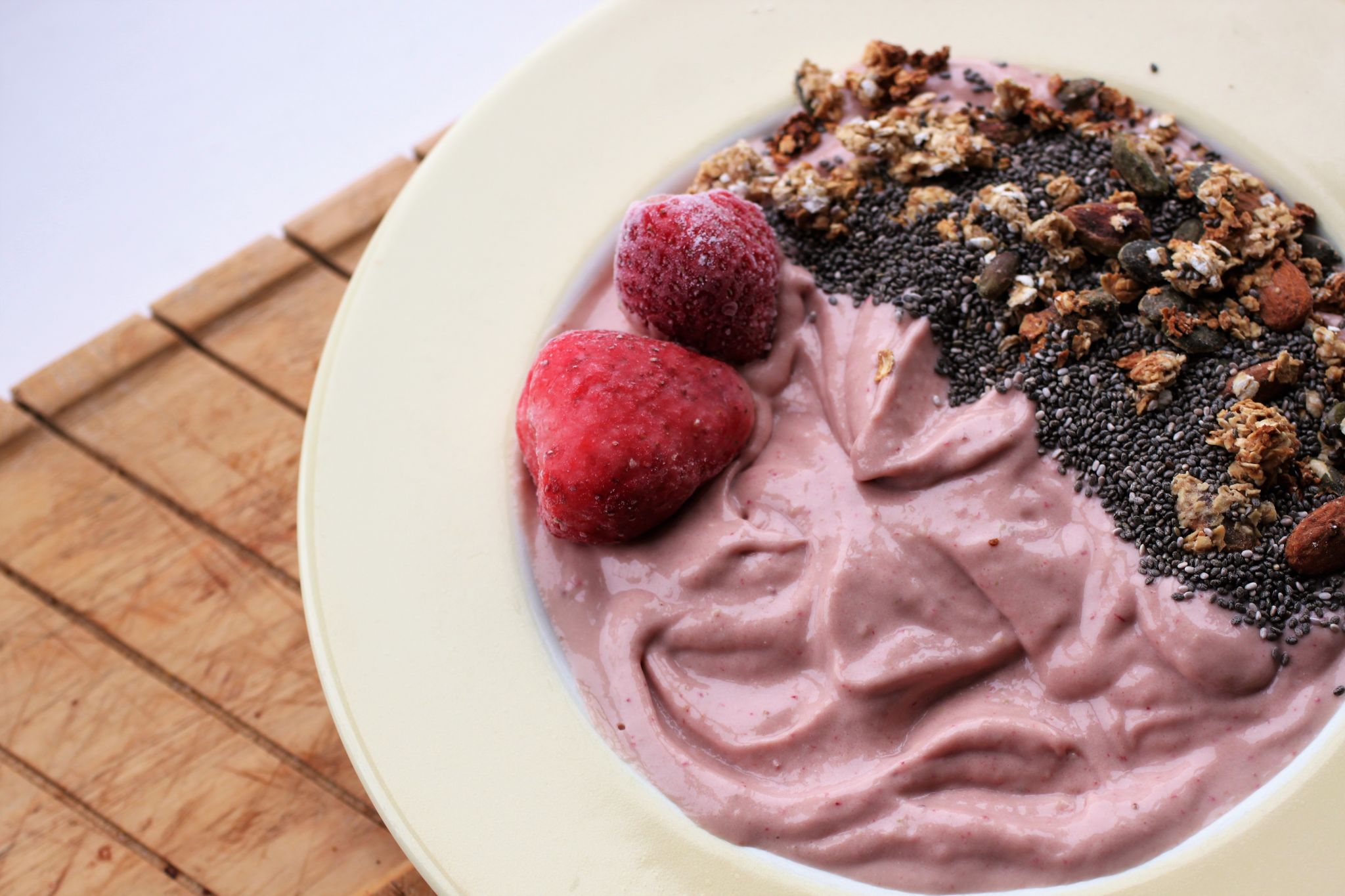 strawberries and cream smoothie bowl in a light yellow bowl, sat on a wooden board. Picture taken from above, flat shot.