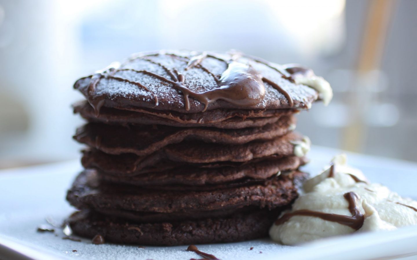 chocolate orange pancakes stacked on a white plate, served with cashew cream and sprinkled with icing sugar