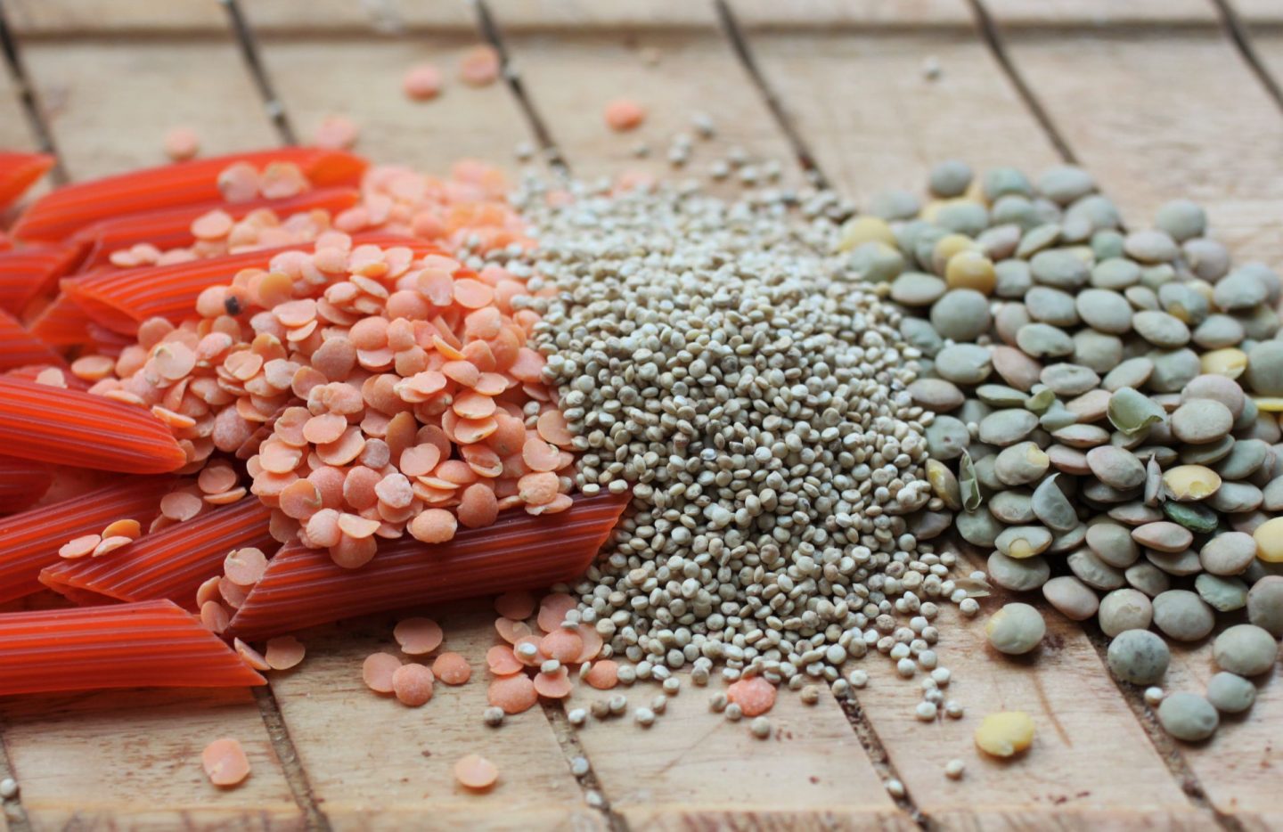 a wooden board with some vegan protein sources spread out on, lentil penne, red lentils, quinoa and green lentils