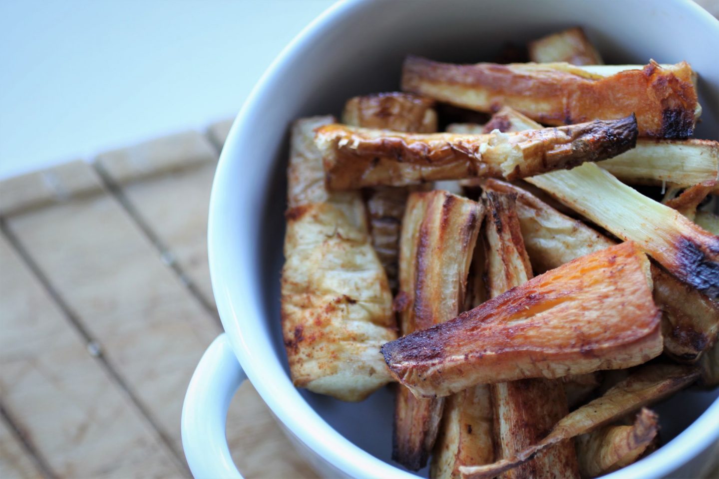 close up shot of a bowl of chilli roasted parsnips in a while bowl, sat on a pale wooden chopping board
