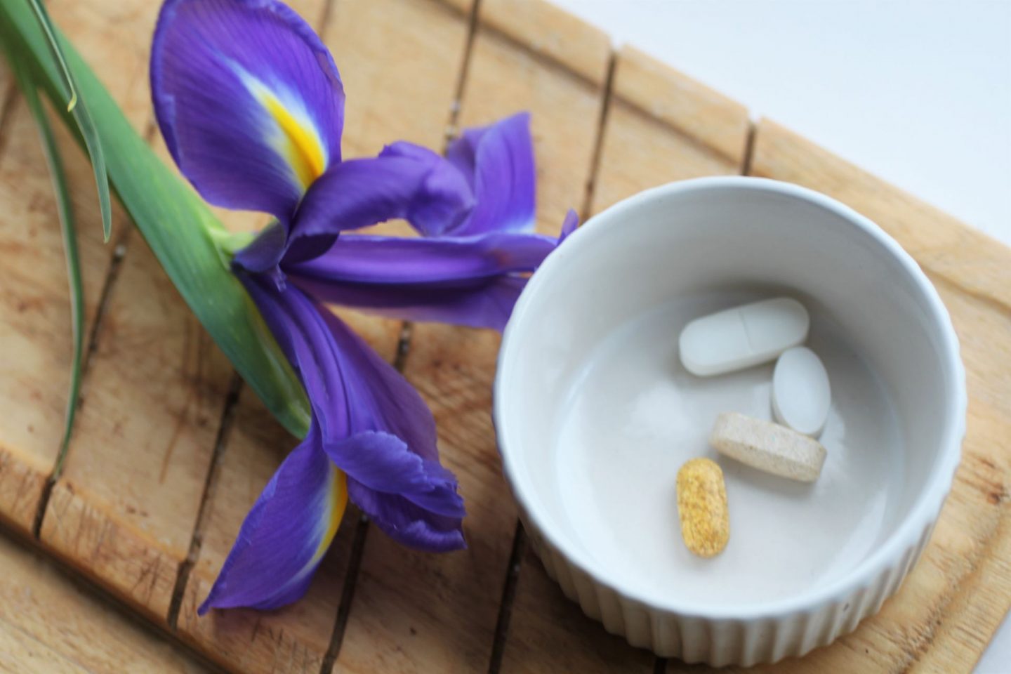 small round white ramakin with 4 pills in it on a wooden board next to an iris