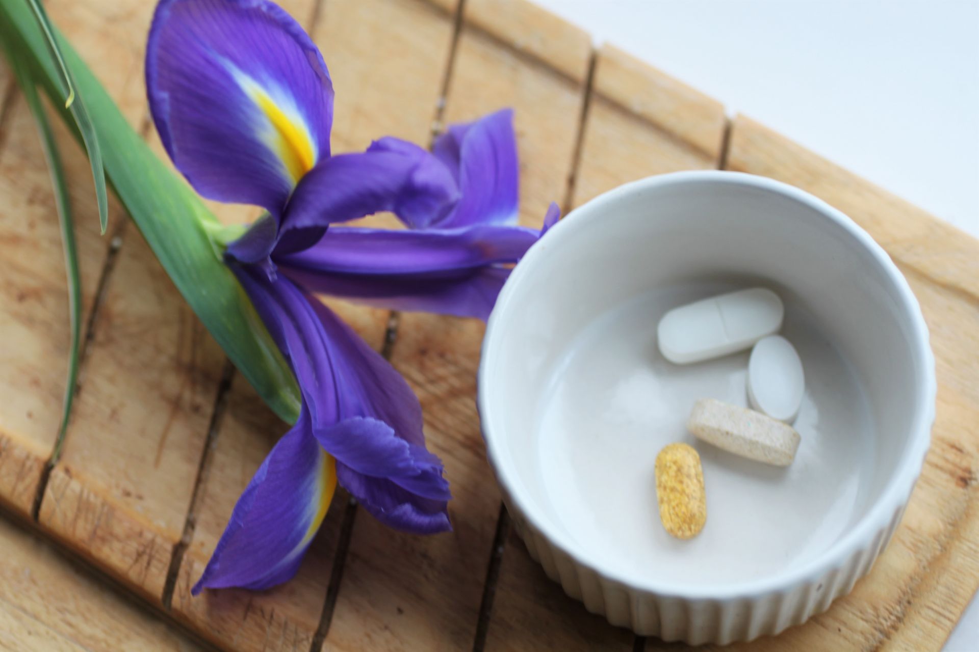 small round white ramakin with 4 pills in it on a wooden board next to an iris