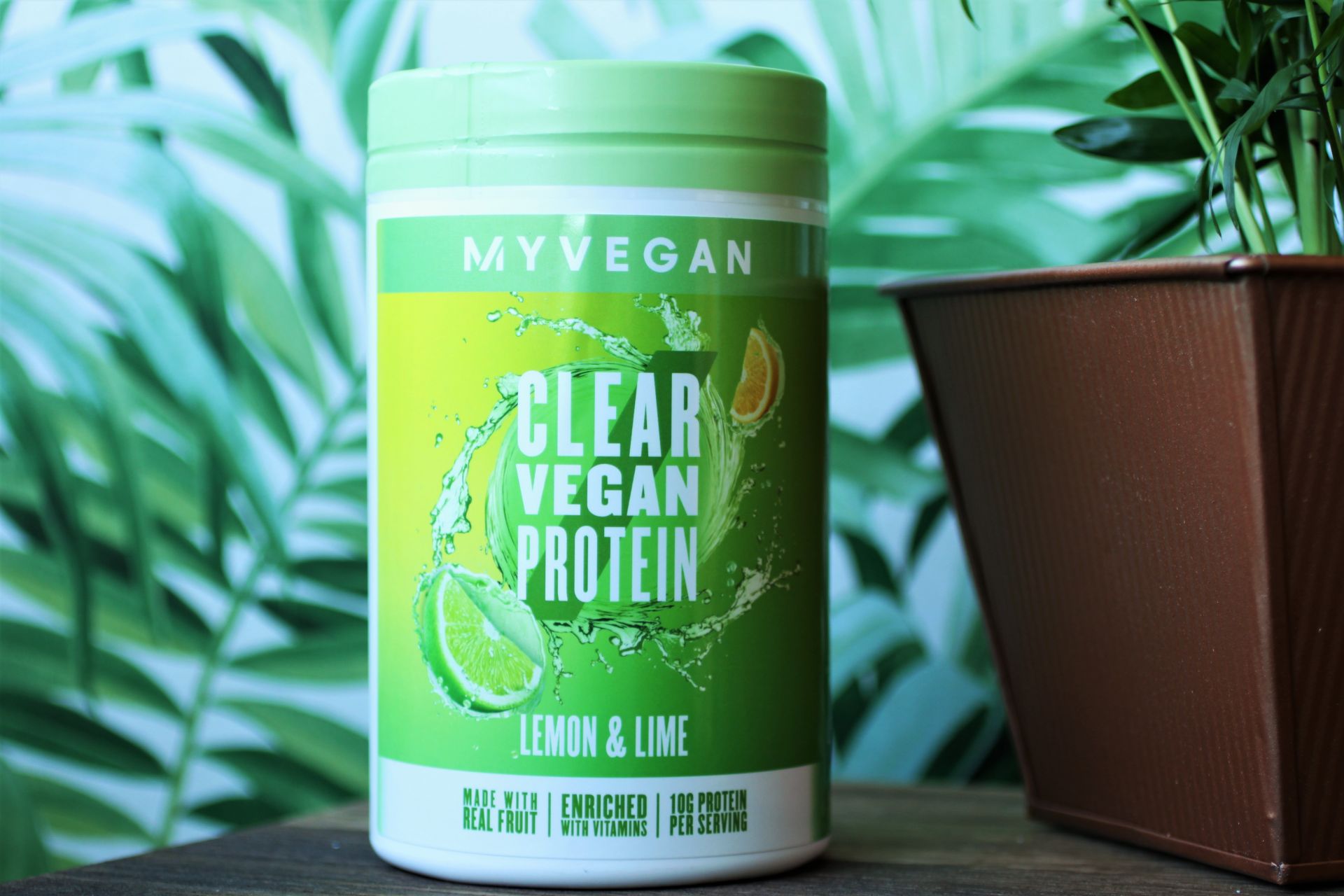 a tub of lemon and lime clear vegan protein sat on an end table, next to a plant in a copper pot with leaf print wallpaper in the background