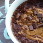 a white bowl on a wooden tray with brownie batter dessert oatmeal inside. Brown oats with a swirl of nut butter in the middle and chocolate chips