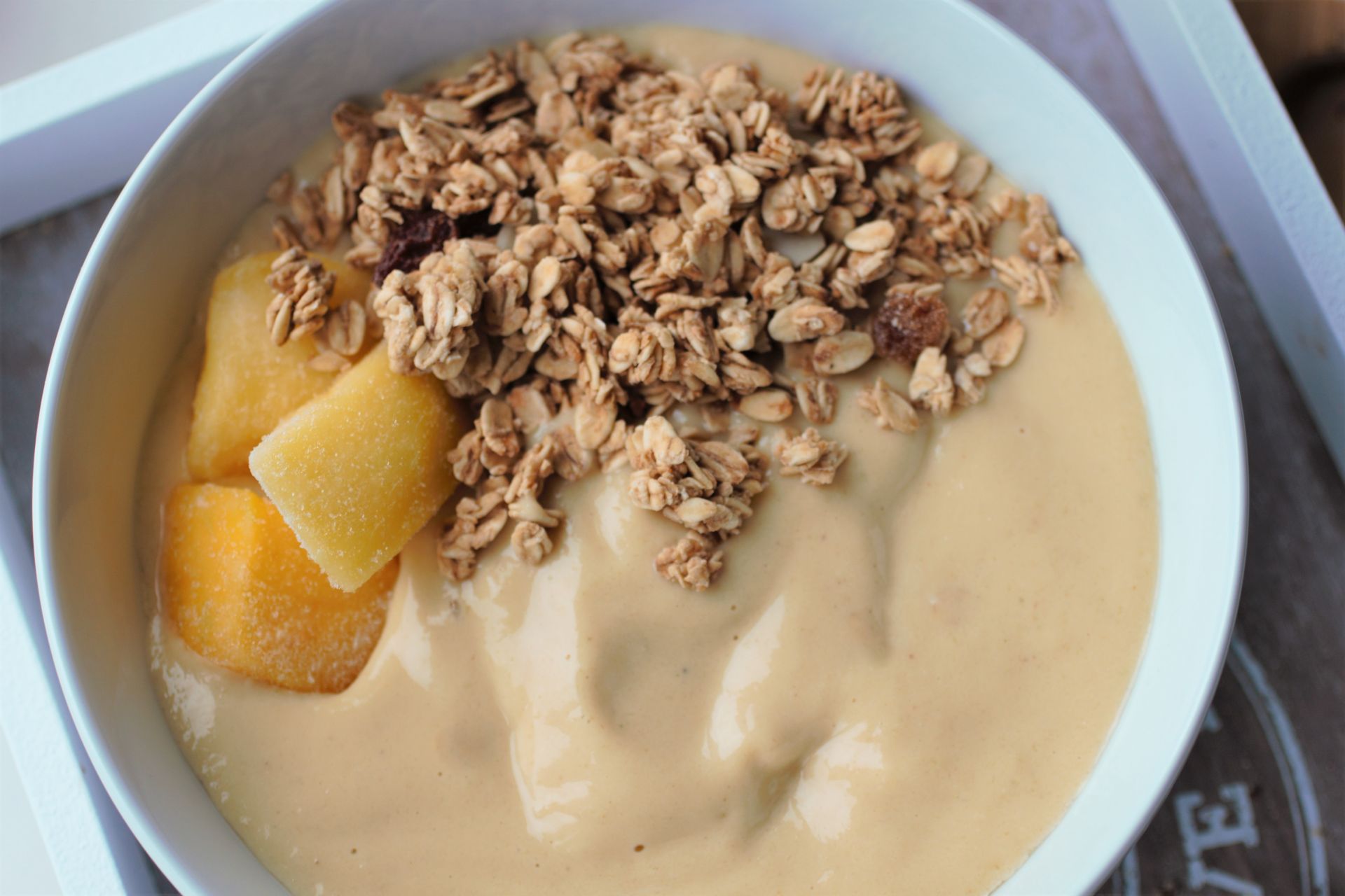 tropical smoothie bowl topped with granola and some mango chunks. In a white bowl placed on a tray with a white wooden rim