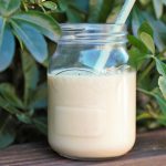 tropical smoothie in a kilner jar, placed on a wooden bench with green leaves/ bushed behind it