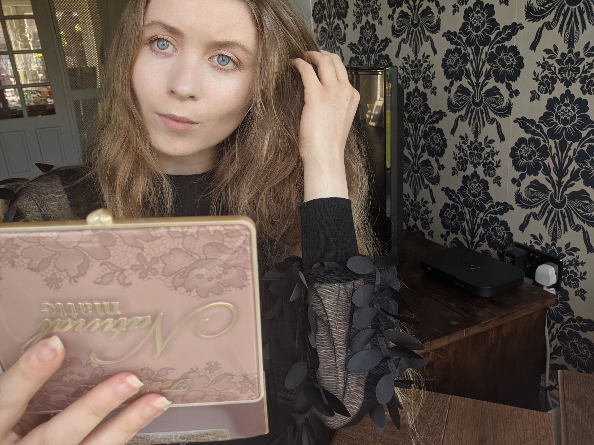 GRWM coronavirus style, me with my makeup nearly done, holding up a too faced palette mirror to check my progress