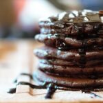 a stack of mint chocolate pancakes on a wooden tray, topped with icing sugar and chocolate sauce