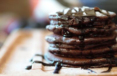 a stack of mint chocolate pancakes on a wooden tray, topped with icing sugar and chocolate sauce