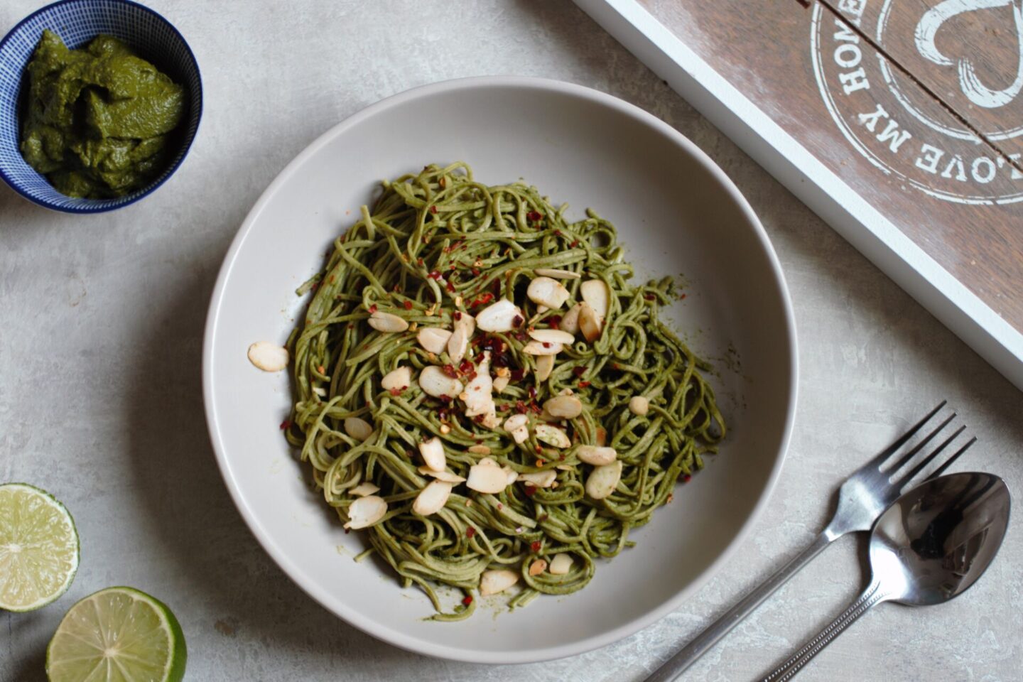 a bowl of edamame spaghetti against a grey background with a sliced lime and pot of pesto beside it