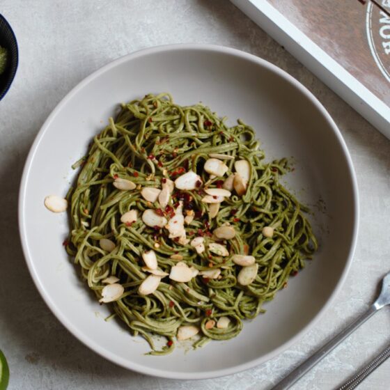 a bowl of edamame spaghetti against a grey background with a sliced lime and pot of pesto beside it