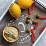 a small pot of lemon & olive pesto on a grey tray with olives and pasta scattered around it