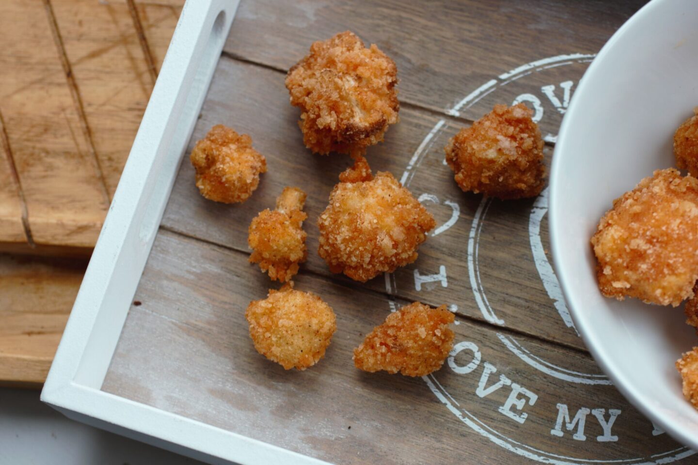 wooden tray with popcorn cauliflower pieces on it with a white bowl filled with popcorn cauliflower to the right edge of the frame