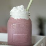 cherry bakwell milkshake recipe in glass jar topped with vegan cream, placed on a wooden tray with a blurred background