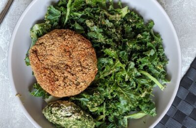 spinach burgers recipe - one spinach burger and half a burger without breadcrumbs in a grey bowl, on a bed of greens