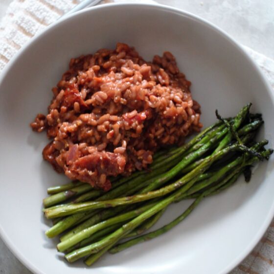 a bowl of red wine risotto with garlic butter asparagus in a stone bowl placed on a neutral check tea towel