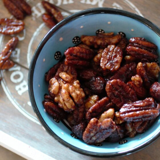 a bowl of the orange candied nuts recipe with a few nuts sprinkled on a wooden tray in the background