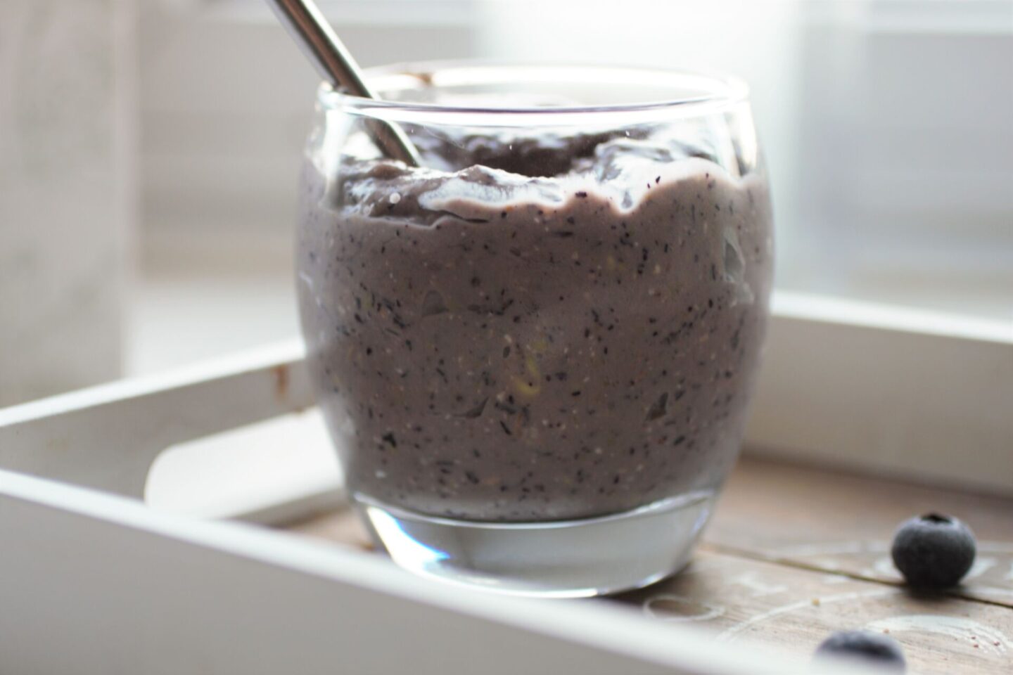 a glass of blueberry breakfast pudding placed on a tray with some blueberries scattered around