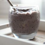 a glass of blueberry breakfast pudding placed on a tray with some blueberries scattered around