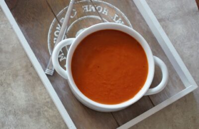 a serving of carrot & red pepper soup in a white bowl placed on a grey wooden tray