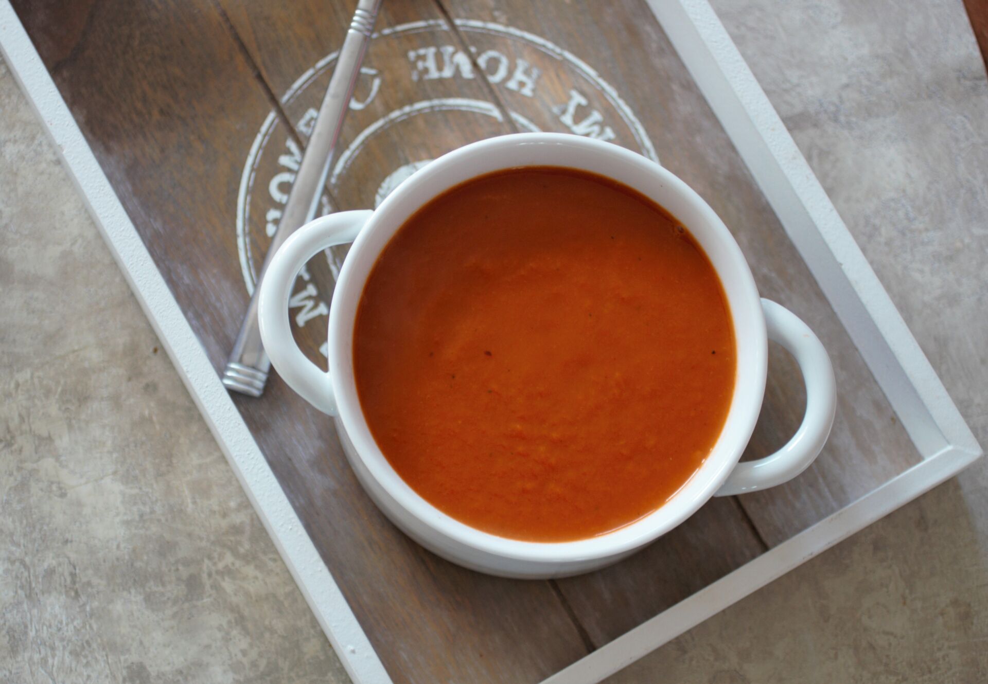 a serving of carrot & red pepper soup in a white bowl placed on a grey wooden tray
