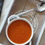 a bowl of carrot & red pepper soup placed on a wooden tray with a spoon next to it