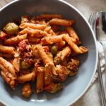 bowl of sundried tomato pasta bake, placed next to a fork and spoon, on a grey background, shot from above