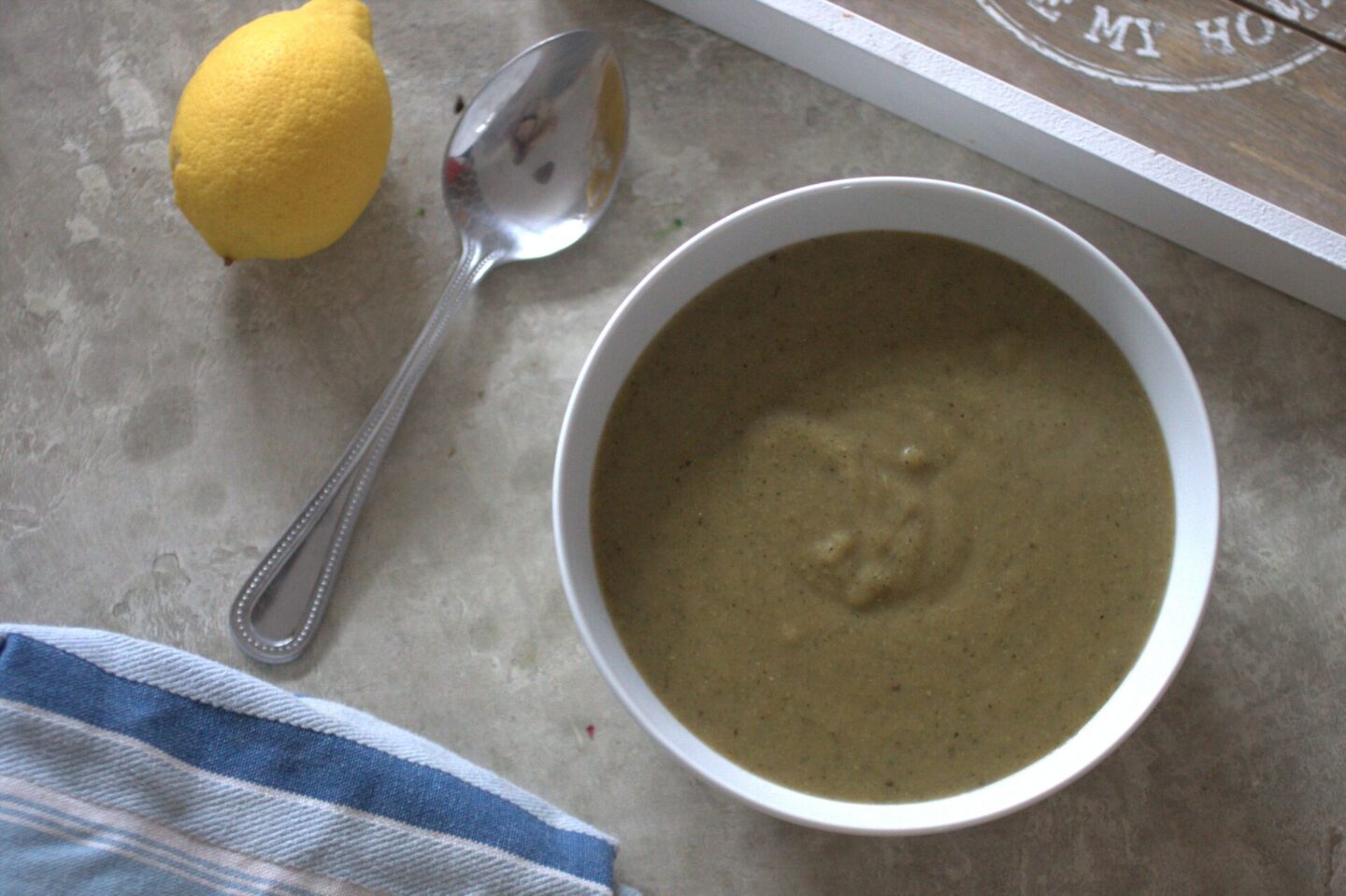 a bowl of broccoli soup recipe on a table, shot from above with a tea towel, lemon and spoon placed around it