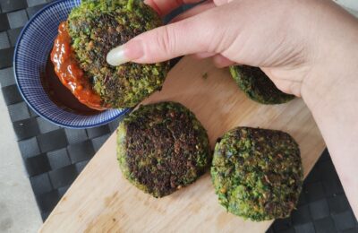 Wooden board with broccoli and kale fritters on it, with one being dipped into a pot of hot sauce