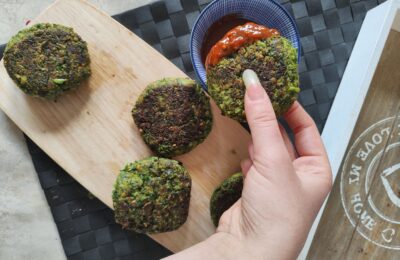 Wooden board with broccoli and kale fritters on it, with one being dipped into a pot of hot sauce