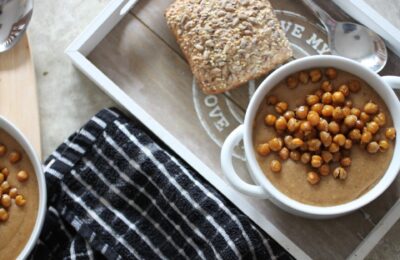 two bowls of spiced cauliflower soup on a dining table, one bowl placed on a tray with a bread roll