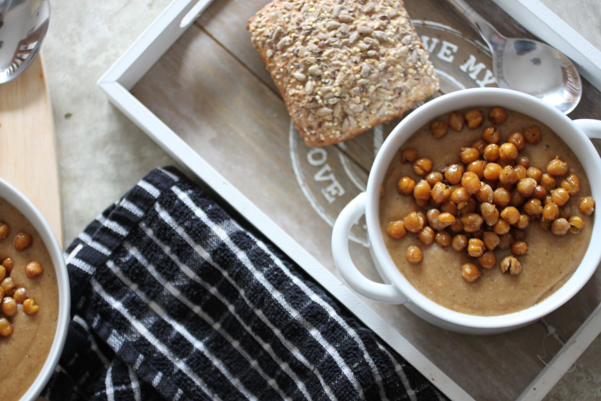 two bowls of spiced cauliflower soup on a dining table, one bowl placed on a tray with a bread roll