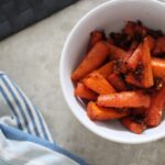 harissa parm carrots in a white bowl placed on a table next to a blue striped tea towel