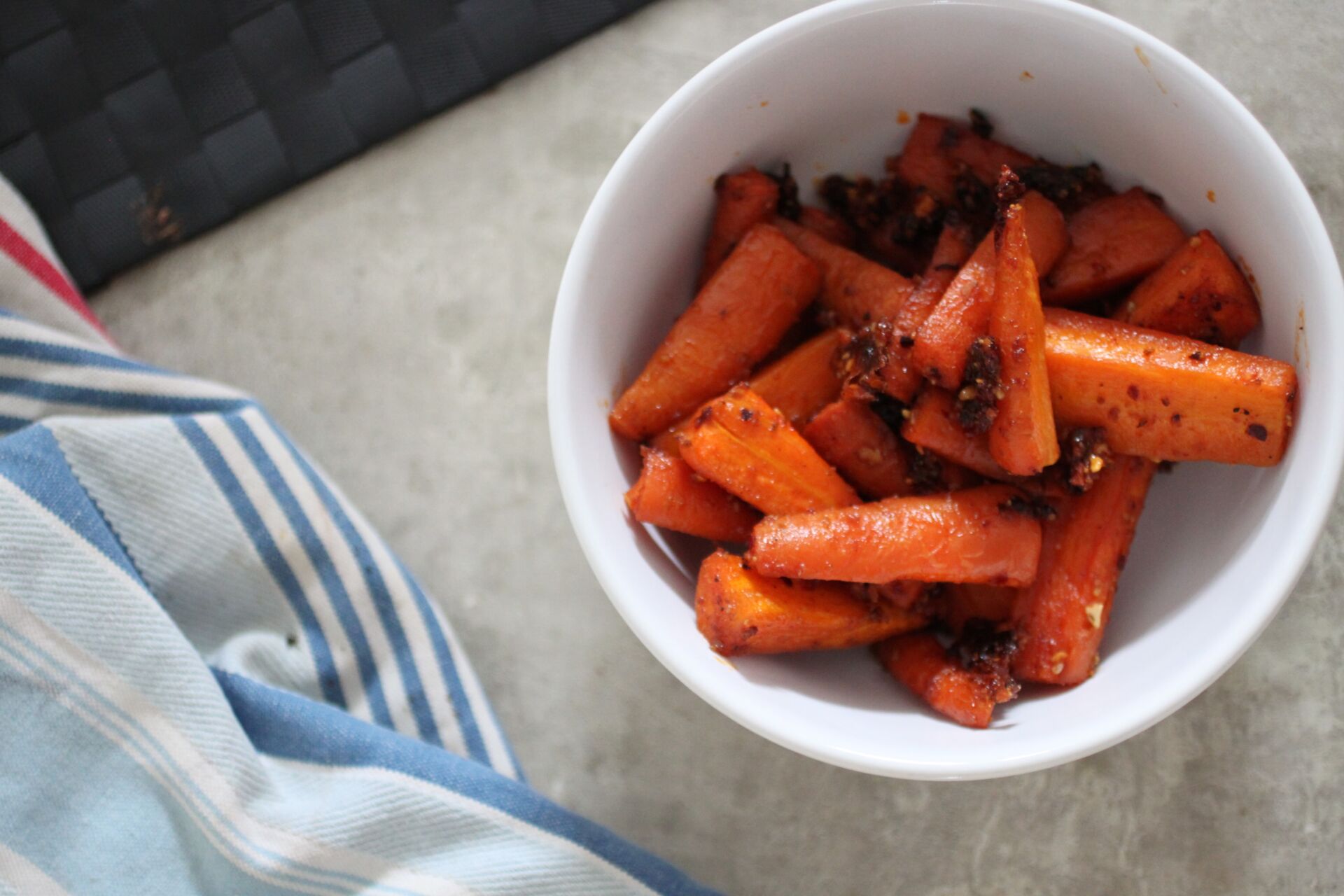 harissa parm carrots in a white bowl placed on a table next to a blue striped tea towel