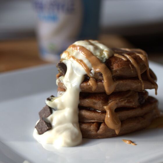 Vegan GF peanut butter protein pancakes with toppings, on a white plate with peanut butter dripping down