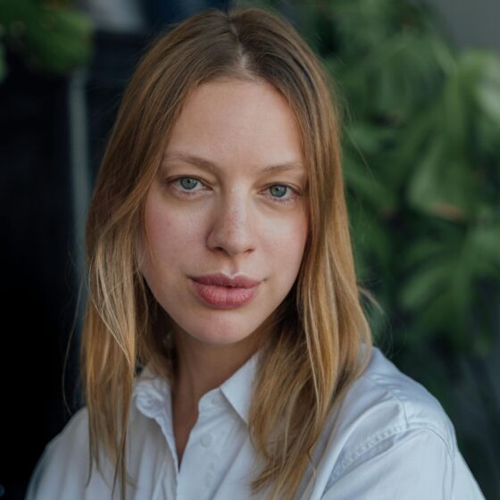 woman with fuller lips, blonde hair and grey eyes standing in front of plants