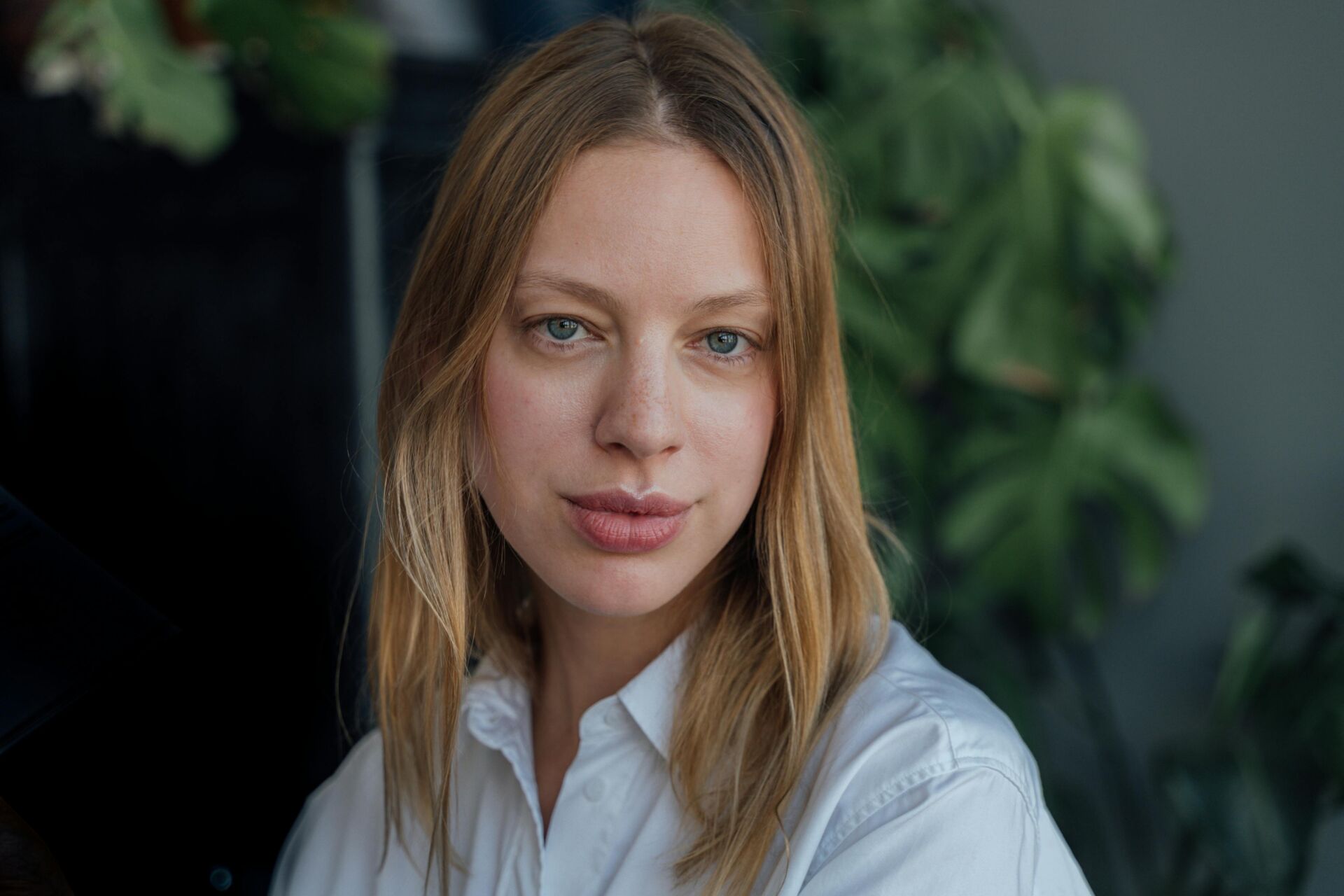 woman with fuller lips, blonde hair and grey eyes standing in front of plants